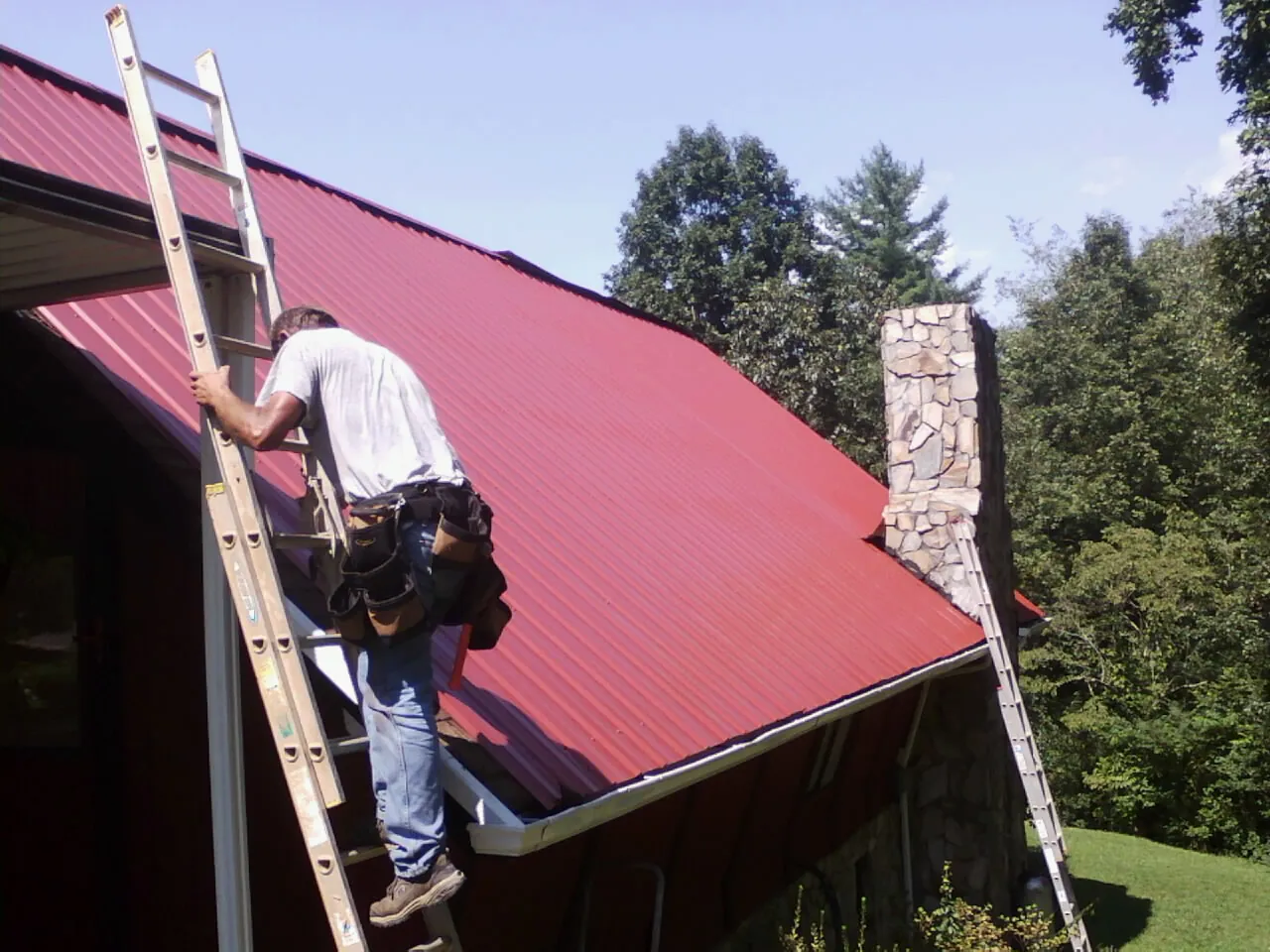 Roofer on a ladder beside a red metal roof and stone chimney — Soffit & Fascia Repair by Roofing Brothers in Canon City